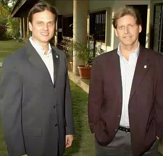 Dos hombres de traje posando juntos al aire libre, frente a un edificio